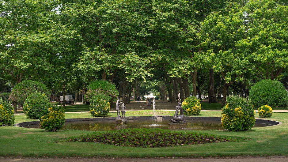A landscaped garden scene featuring a circular fountain with three metal sculptures of children playing, surrounded by neatly trimmed, rounded green shrubbery with yellow-tinted foliage. The fountain 