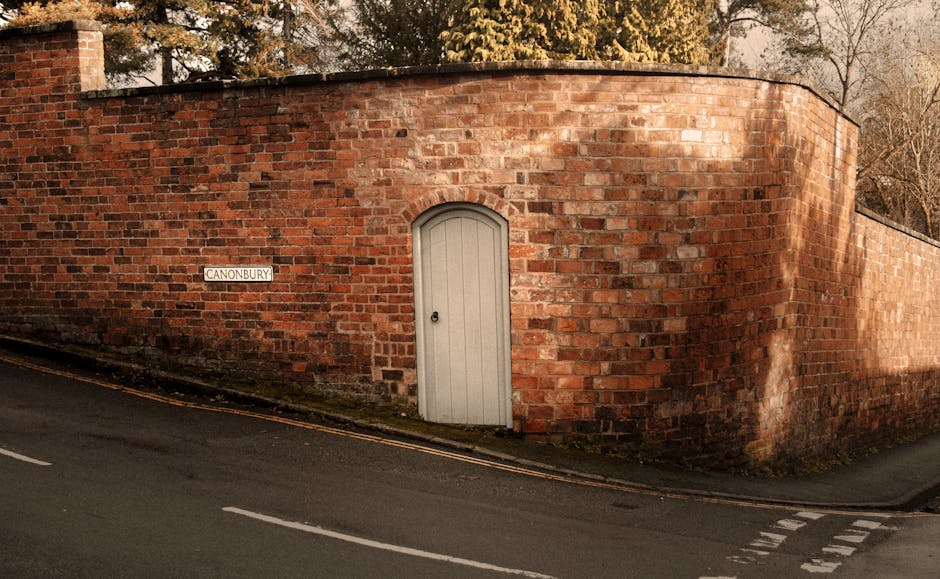 A curved red brick wall with a textured surface surrounds a garden area, featuring a narrow white door with an arched top set into the wall. To the left, a small white street sign reading 'CANONBURY' 
